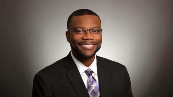 Dr. Winston McCowan poses for a headshot against a vignette background wearing a dark suit, purple-toned tie and a gold cross necklace.